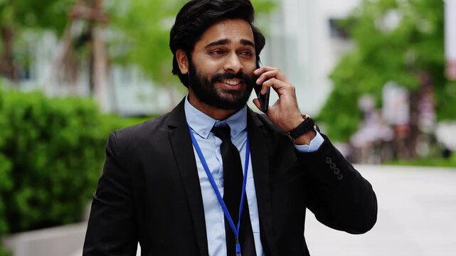 Indian businessman walks along a city walkway while talking on his smartphone, his expression turning worried and serious as the camera moves in close to his bearded face