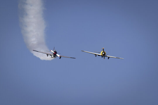 View of two vintage propeller aircraft performing an aerobatic maneuver with smoke trails against a clear blue sky in La Spezia, Liguria, Italy.