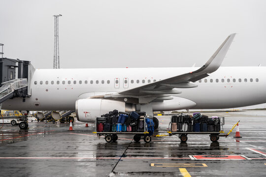 View of a white passenger airplane parked at an airport gate with luggage carts loaded with suitcases on the wet tarmac in Iceland.