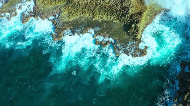 Aerial view of turquoise ocean waves crashing against rugged rocky