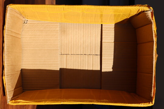 Open cardboard box sealed with yellow packing tape, viewed from above. Strong sunlight creates geometric shadows inside the empty box, highlighting its rough texture and handmade appearance.