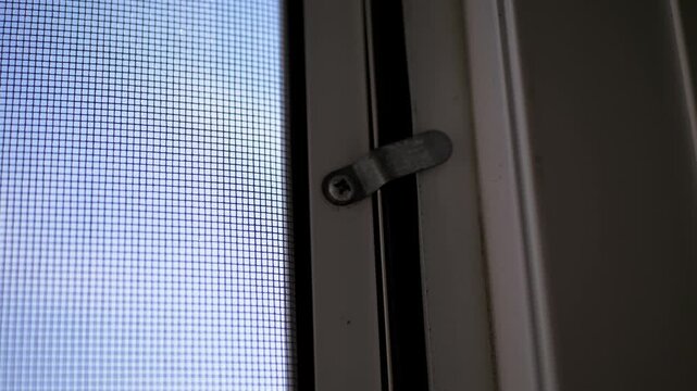 Metal swivel clip firmly secures a fine mesh insect screen against a white window frame. Daylight filters through the tiny square apertures, highlighting functional residential hardware.