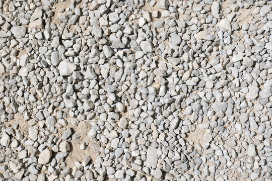 Grey crushed stone gravel texture background with sandy soil, small pebbles and dry grass stems, full frame top view of rough aggregate surface, natural construction material detail