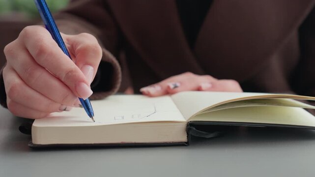 Close up shows woman hands holding blue pen drawing simple geometric shapes in open notebook on table. focus remains steady capturing detailed hand movements and paper texture