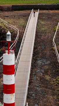 Woman walking toward lighthouse in Azores, Portugal. Vertical aerial footage of solo female traveler on wooden boardwalk across volcanic terrain