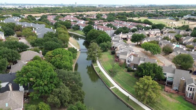Overhead view follows meandering canal channel dividing compact suburban residential lots. Drone perspective showcases tree canopies arching over paved walkways in, Valley Ranch, Irving, TX