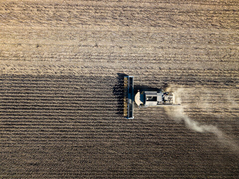 Agricultural tractor plows soil for sunflower cultivation, carving parallel tracks through broad field. Dust rises behind farm vehicle sunflower plants are not visible in this overhead view.