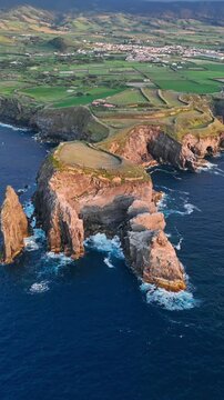 Aerial view of dramatic volcanic cliffs and sea stacks along a rugged coastline with green fields and a distant village