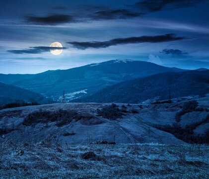 mountainous rural landscape of ukraine on spring night. picturesque highland under dark sky. grassy meadow in full moon light. carpathian countryside scenery with forest on a hill behind the pasture