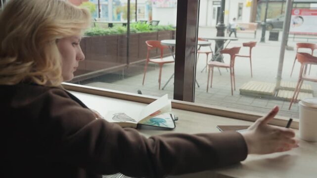 Woman sketches in cafe at table with drawing tools. Woman holds pencils while seated near art supplies like color palettes and notebooks visible through large windows
