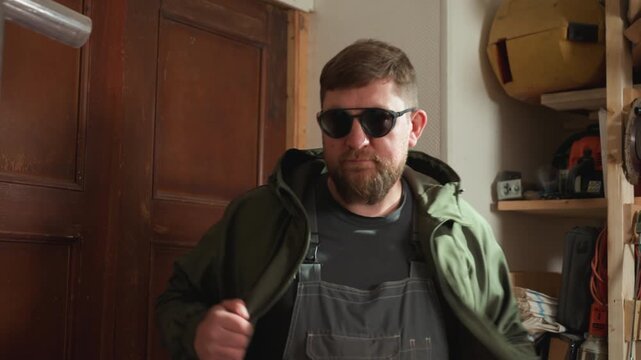 Man removes his green jacket and reveals workwear inside woodworking shop. Man examines door frame while surrounded by tools like saws clamps wood planks hardware measuring tape square