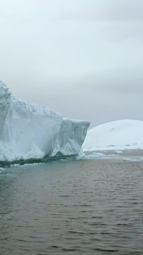 Vertical Video of Iceberg Foreground With Snow-Covered Ridge Behind. White ice, rippling water and snow-covered ridges create a layered cold seascape beneath soft diffuse light.