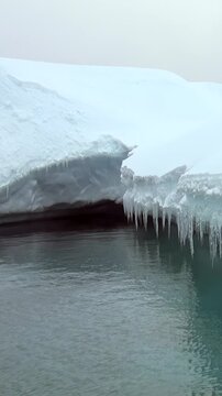 Vertical Video of Curved Low Iceberg Floating on Dark Water. White and blue ice, rippling water and soft diffuse light reveal natural frozen texture and sculpted contours across the marine scene.