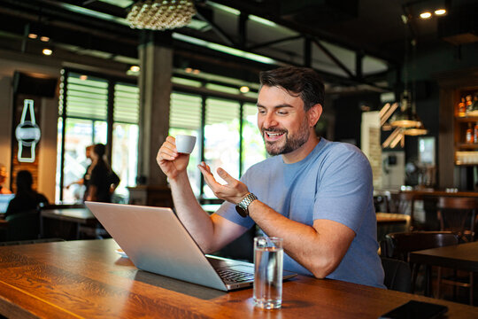 Man drinking coffee and using a laptop for a video call at a cafe bar