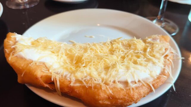 Close-up footage of a Hungarian langos, deep-fried flatbread folded in half and topped with sour cream and grated cheese, served on a white plate in a cafe. High quality 4k footage
