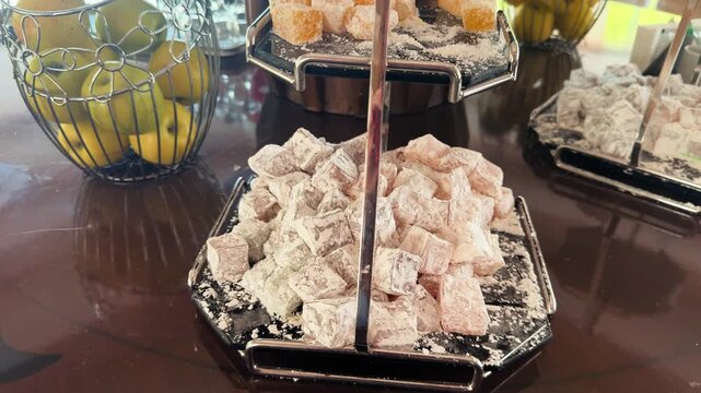 Footage of Turkish delight (lokum) cubes dusted with powdered sugar on a black metal tiered serving stand, traditional oriental sweets on a hotel buffet table, blurred fruit basket in the background