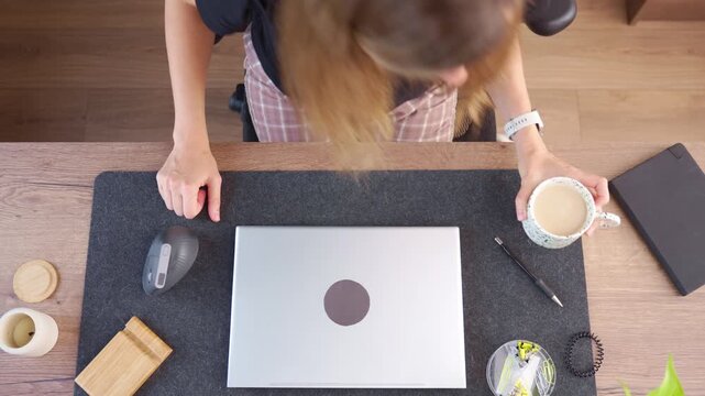Woman approaches desk with coffee, sits down, opens laptop and starts working. Concept of remote work and starting workday at home