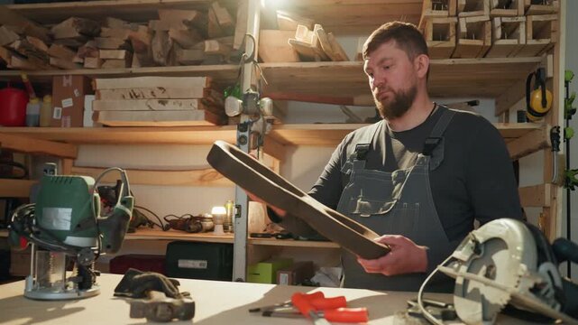 Craftsman inspects shape of handcrafted woodwork at his workshop table. Various power tools like router are visible on shelves with stacked lumber and woodworking materials in background