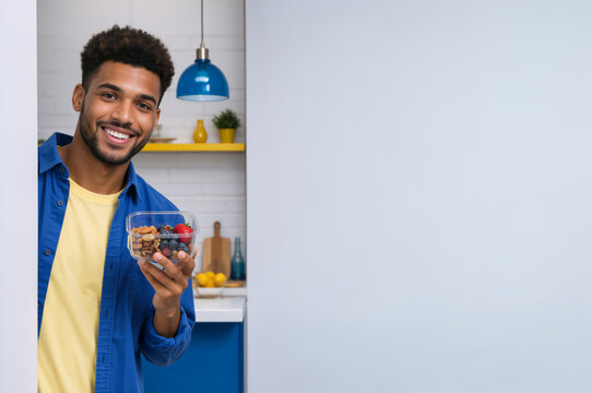 Smiling young man holding a healthy snack container in kitchen. Cheerful adult with fresh berries and nuts. Copy space for text