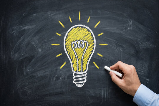 Hand drawing a glowing lightbulb with chalk on a blackboard representing innovation and bright ideas