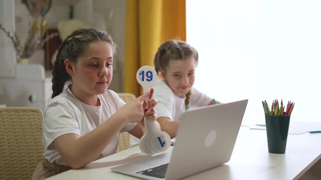 Young girl with braided hair holding math flashcards showing numbers while teaching online. Her sister smiles working on laptop computer. Colorful pencils and notebooks on white study table.