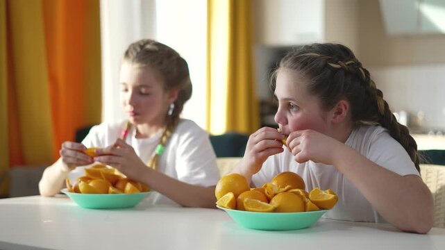 Two girls eating orange slices at home kitchen table. Sisters enjoy citrus fruit snack together. Children share healthy oranges. Kids eat vitamin-rich fruit. Happy girls with fresh orange slice bowl.
