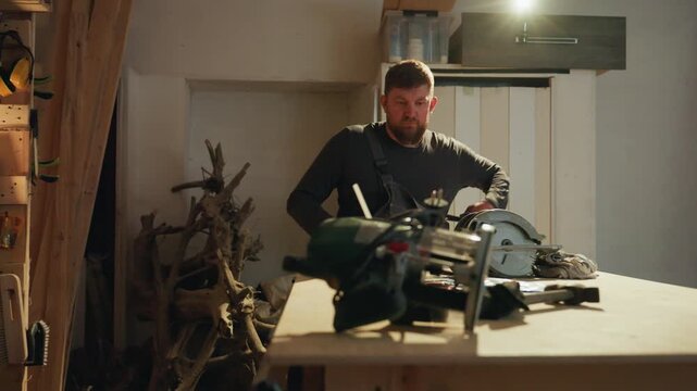 Carpenter organizes safety gear in indoor wood shop surrounded by power tools and lumber. man adjusts his overalls while various woodworking equipment like circular saw clamp ear protection