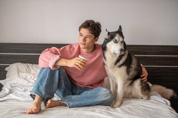 Young owner sitting together with husky on wide bed, coffee drunk beside warm animal, scene that modern brands keep asking for. Wide shot. Concept of morning spent together with own dog © iwavephoto