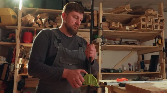 Man in overalls secures wood piece with green clamp on workbench surrounded by woodworking tools and materials including sawdust power tool measuring tape hand tools lumber clamps workshop
