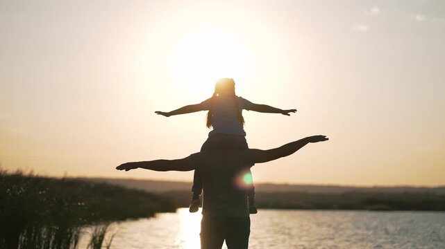 Father carries daughter on shoulders during sunset. Child spreads arms wide like airplane wings by peaceful lake. Parent and kid enjoy outdoor bonding moment in beautiful nature at golden hour.