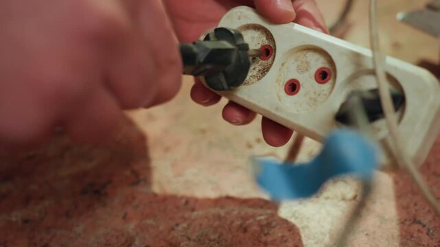 Hand holds dirty power strip with multiple sockets using tools to manipulate plug and socket. focus is on close up details of hands working directly with electrical components and repair
