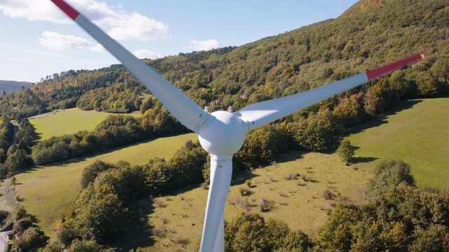 A wind turbine stands tall in a green field surrounded by hills and trees. The scene shows a clear sky and natural surroundings.