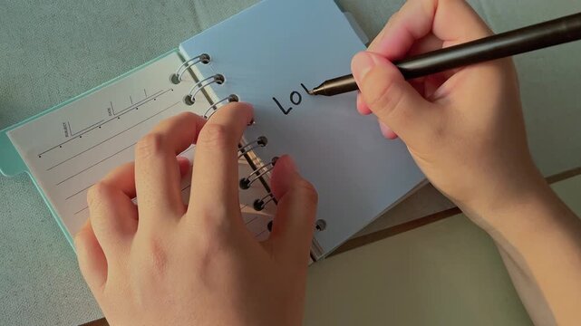 Woman hands writes "Love me" in a small notebook. Soft lighting highlights gentle handwriting. Person holds pen with calm focus. Notebook has lined pages and metal rings. Ideal for romantic