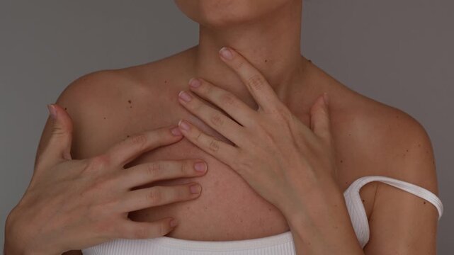 Young woman shows red irritated skin on her chest and collarbone area, gently touching rash and sensitivity against gray background