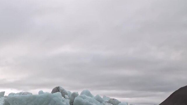 Rugged mountains rise above cold water scattered with floating ice. The frame preserves a cloudy sky as supporting detail, adding natural texture and depth to the view.
