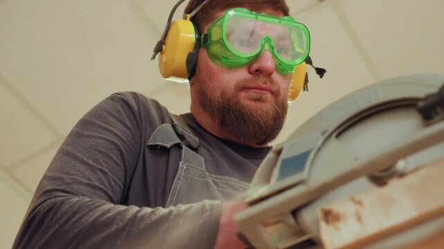 Man wearing green safety goggles and yellow ear protection operates large industrial power tool guiding material through machine. Dust particles visible around working area. Indoor setting