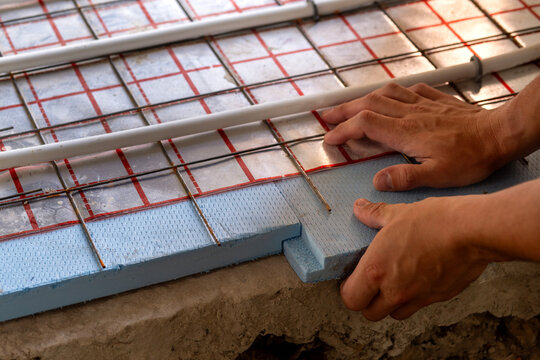 Closeup of contractor hands arranging plastic floor heating pipes across foam board insulation, preparing an energy-efficient radiant heating system for a new home.