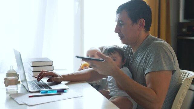 Father multitasking work from home with two babies. Dad works on laptop while making phone call. Parent juggles childcare and remote work. Man balances fatherhood with professional tasks.
