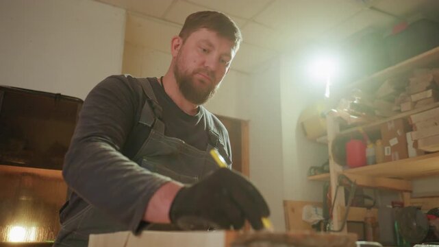 Craftsman wearing dark workwear carefully traces lines on raw lumber in his workshop. scene includes various woodworking tools and materials with warm lighting illuminating workspace