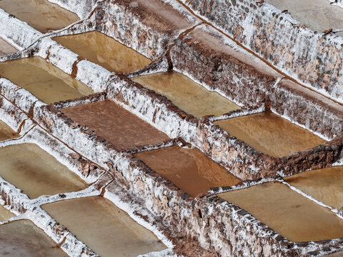 Terraced salt evaporation ponds with white crystallized salt walls and brown water seen from above at the Maras salt mines, Cusco