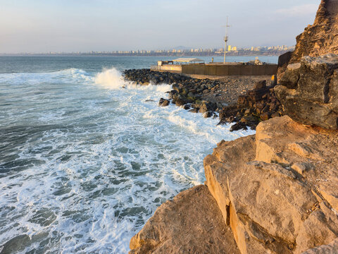 Waves crashing against cliff rocks on the coast with a breakwater in the background and a coastal city on the horizon