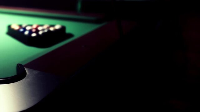 A pool table is seen in a dark room with balls arranged in a triangle. The game is about to start as players gather around the table ready to play.