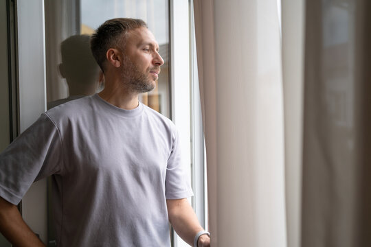 Confident man by window enjoys quiet optimism, relief and gratitude after personal success, thinking about future with calm satisfaction, renewed motivation and positive outlook.