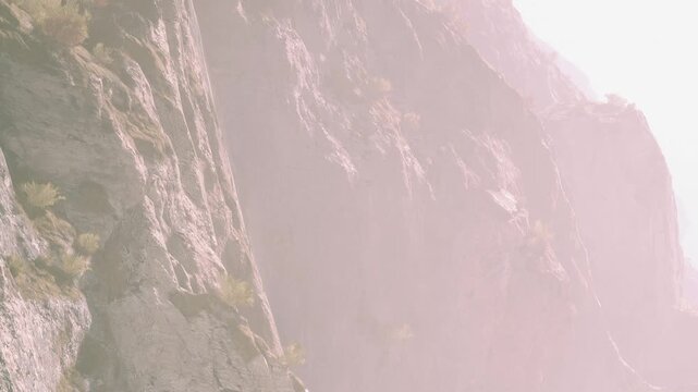 People are climbing steep cliffs in a rocky mountain area. The sun shines brightly, casting shadows on the rocks. Green plants grow on some cliff sides, showing life in the rough terrain.
