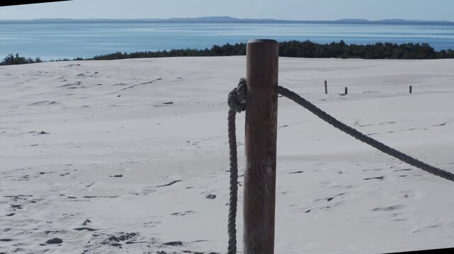 Wooden post and rope fence in a vast sandy coastal landscape, with calm blue water and distant hills. The scene evokes boundaries, freedom, solitude, and escape.