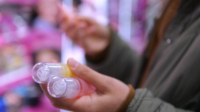 Woman holds two skincare bottles while browsing beauty store. Shopper examines clear cosmetic containers and compares options among softly blurred shelves of products, considering product selection.