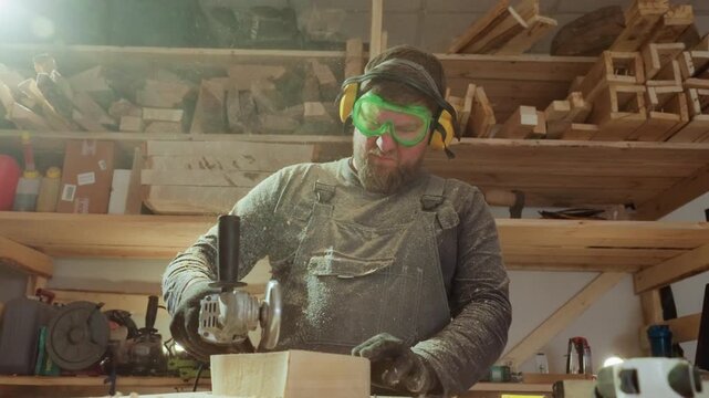 Carpenter uses angle grinder on wooden material within well equipped woodworking shop. man wears safety goggles and ear protection while operating power tool with wood planks visible