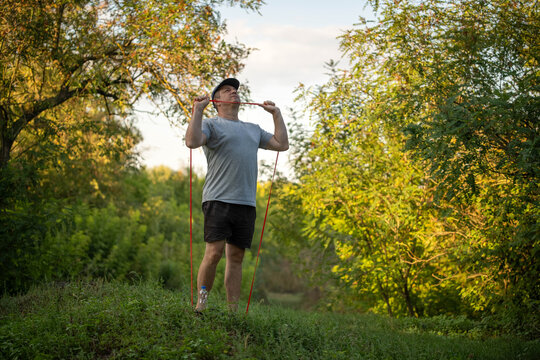 Man performs resistance band stretch in forest clearing, lifting red cord beside shoulders. Focused exerciser stands on grassy path amid sunlit trees, doing controlled fitness training outdoors.