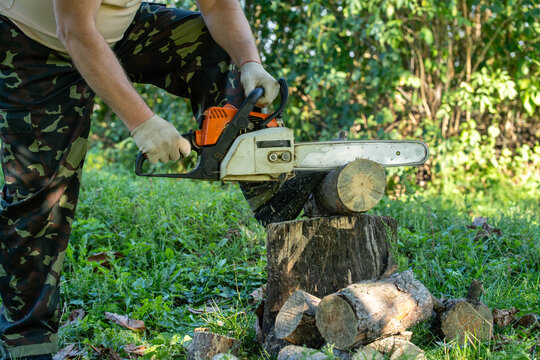 Woodcutter holds chainsaw to cut log resting on stump in green forest clearing. Man wears work gloves and camouflage trousers while preparing firewood among cut timber and grass, gripping saw handle.