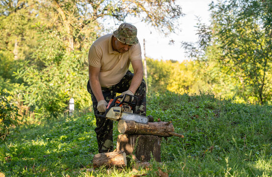 Man operates chainsaw on log in woodland. Attentive worker wears camouflage clothing and gloves while preparing firewood on tree stump amid green undergrowth, cutting through trunk with powered saw.